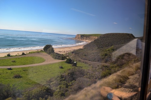 Scenic coastal view with sandy beach, green hills, and calm blue ocean under a clear sky, seen from a train window.