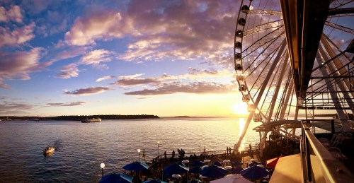A sunset view over water with a Ferris wheel and people enjoying the scene on a waterfront promenade.