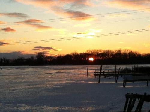 Sunset over a frozen lake, with silhouettes of trees and a dock in the foreground against a colorful sky.