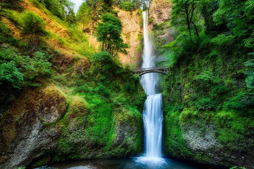 A stunning waterfall cascades down rocky cliffs, surrounded by lush greenery and a bridge above the falls.