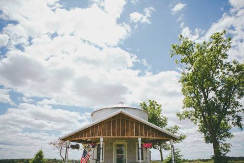 A charming round house with a porch, surrounded by trees and a blue sky filled with fluffy clouds.