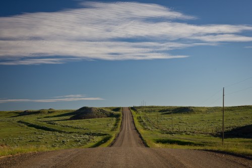 A long, winding dirt road stretches through green fields under a blue sky with wispy clouds.