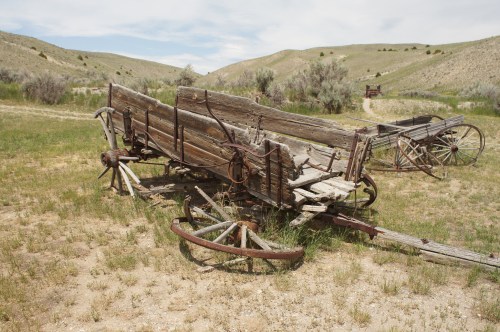 An old, weathered wooden wagon sits in a grassy field, surrounded by rolling hills and sparse vegetation.