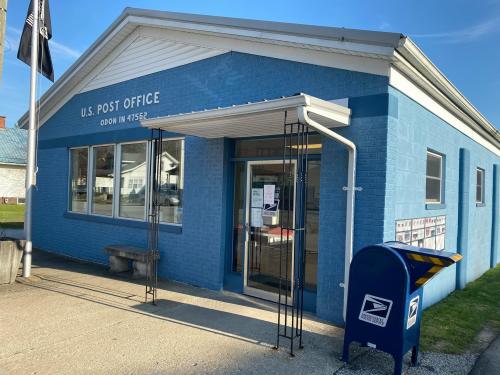 A blue U.S. Post Office building with a mailbox outside, located in Odon, Indiana. Clear sky in the background.