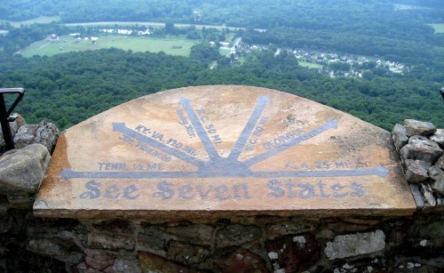 A stone marker on a cliff edge, indicating distances to seven states with a scenic view of green valleys below.
