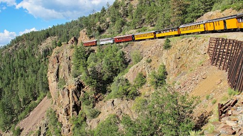 A colorful train winds along a rocky cliff surrounded by lush green trees and a blue sky with scattered clouds.