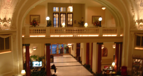 Elegant hotel lobby with high ceilings, ornate decor, and warm lighting, featuring a balcony and lush plants.