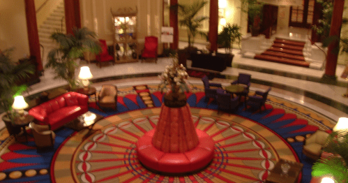 Luxurious hotel lobby featuring a circular carpet, red seating, and tropical plants in an elegant setting.