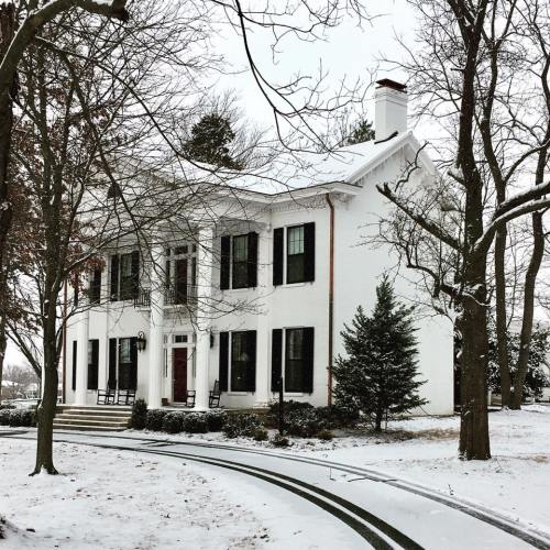A white two-story house with black shutters surrounded by snow-covered trees and a winding path.