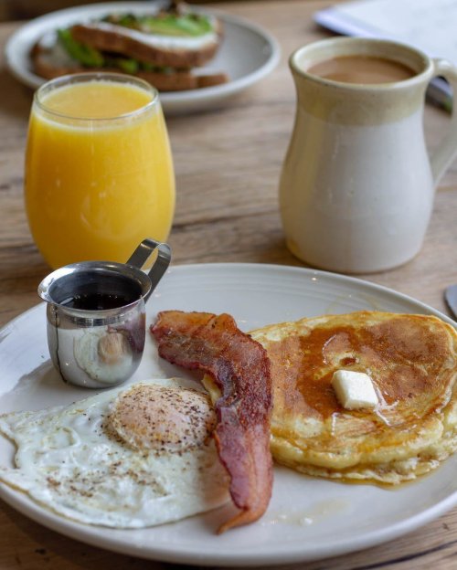A plate with pancakes, bacon, a fried egg, and syrup, accompanied by orange juice and coffee in the background.