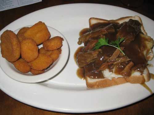 A plate with sliced meat in gravy on bread, served with a side of golden fried croquettes.