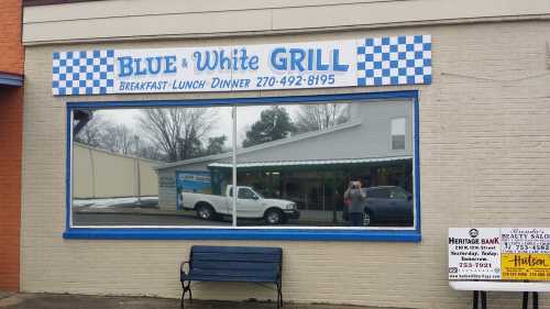 Exterior of Blue & White Grill with a checkered sign, reflecting a truck in the window and a bench in front.