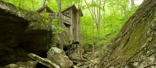 A wooden cabin nestled among lush green trees and large rocks in a serene forest setting.