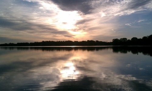 A serene lake at sunset, with colorful clouds reflecting on the water's surface and a silhouette of trees in the background.