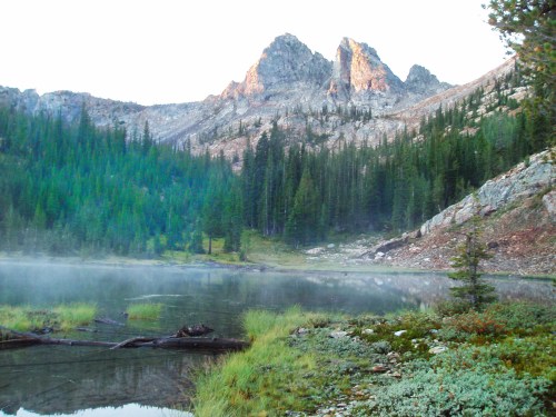 Serene mountain landscape with a misty lake, surrounded by lush greenery and rocky peaks in the background.