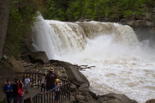A waterfall cascades over rocks, with visitors observing from a nearby path surrounded by lush greenery.