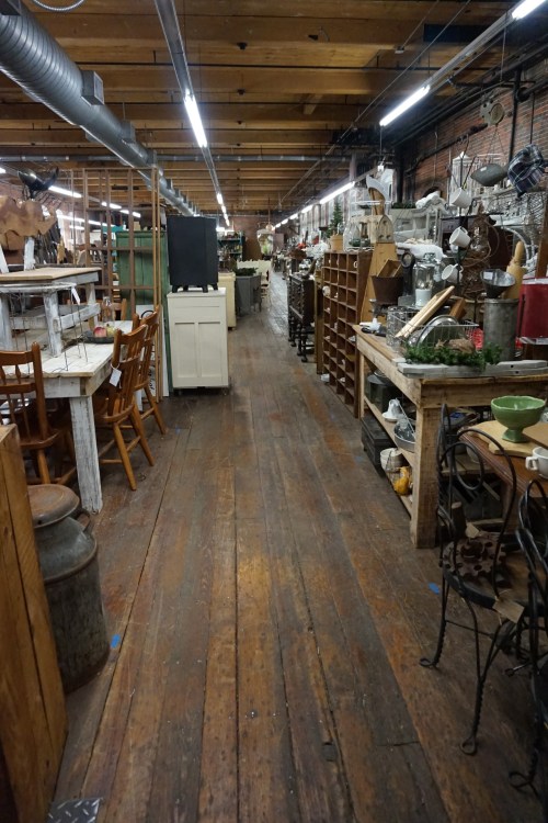 A spacious vintage shop aisle lined with wooden furniture, decor items, and rustic shelves under bright overhead lights.