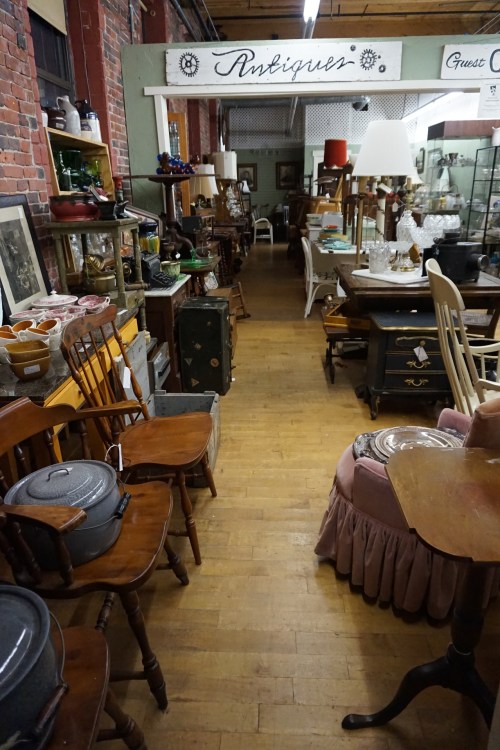 A vintage shop aisle filled with various antique furniture and decor items, including chairs, tables, and glassware.