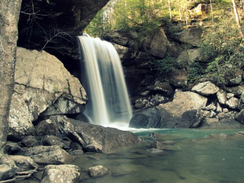 A serene waterfall cascades over rocks into a clear pool, surrounded by lush greenery and rugged cliffs.