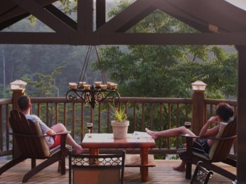 Two people relax on a deck, enjoying drinks and a scenic view of trees in the background.