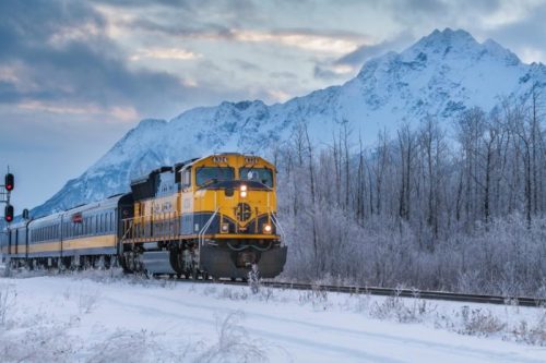 A yellow and black train travels through a snowy landscape with mountains in the background and trees covered in frost.