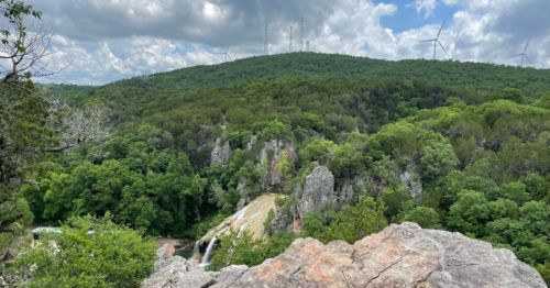 A scenic view of a lush green landscape with a waterfall, rocky outcrops, and wind turbines in the background.