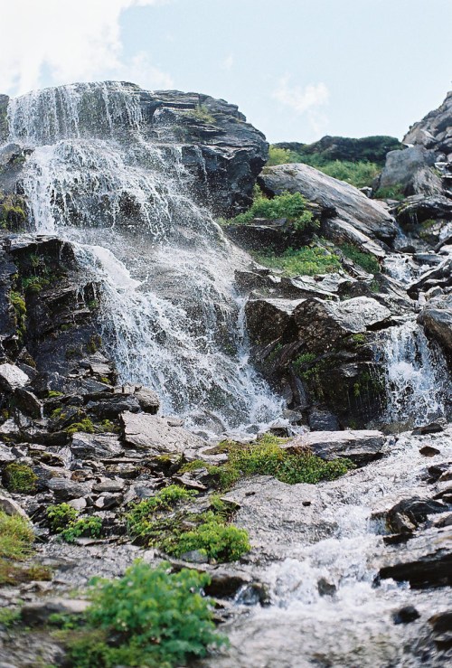 A cascading waterfall flows over rocky terrain, surrounded by lush greenery under a clear blue sky.