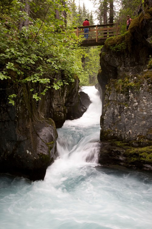 A scenic view of a rushing river flowing through rocky cliffs, with a wooden bridge and lush greenery surrounding it.