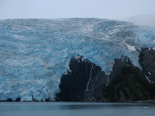 A large glacier with blue ice and rocky cliffs, reflecting in calm waters under a cloudy sky.