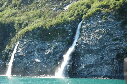 Two waterfalls cascading down a rocky cliff into a turquoise body of water, surrounded by lush greenery.