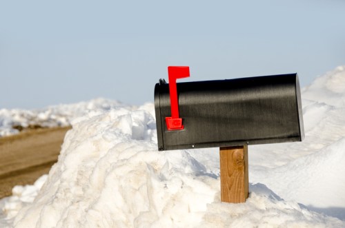 A black mailbox with a red flag stands on a wooden post, surrounded by snow on a clear day.