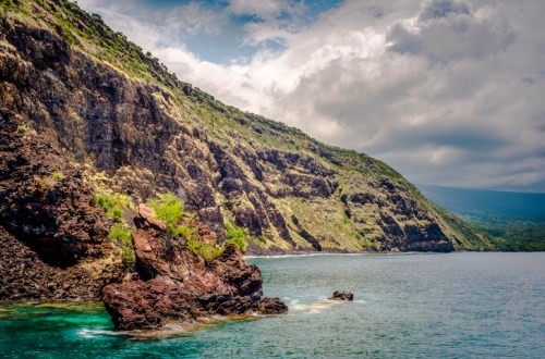 A rocky coastline with lush green hills under a cloudy sky, reflecting in calm blue waters.