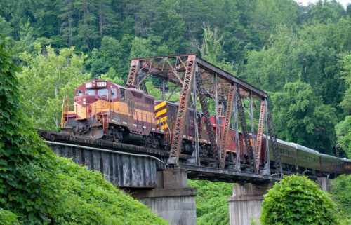 A train crosses a rusted metal bridge surrounded by lush green trees.