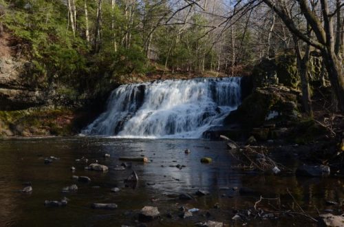 A serene waterfall cascades over rocks into a calm river, surrounded by trees and a peaceful natural landscape.