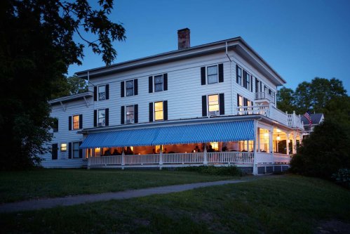 A large white house with blue awnings, illuminated at dusk, surrounded by greenery.