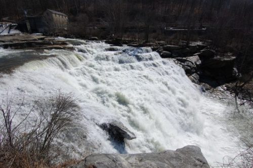 A cascading waterfall flows over rocky terrain, surrounded by trees and a distant building.