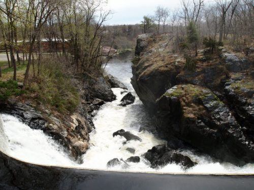 A scenic view of a waterfall cascading over rocks, surrounded by trees and a river in a natural landscape.