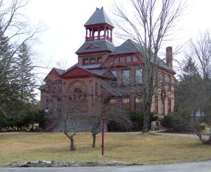 Historic red brick building with a tower, surrounded by trees and a grassy area, under a cloudy sky.