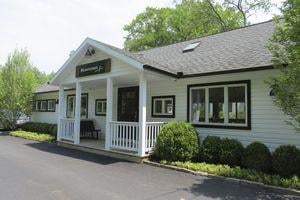 A white, single-story building with a porch, surrounded by greenery and a paved driveway.