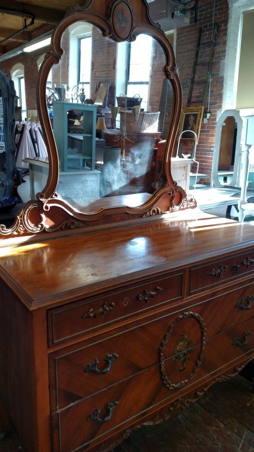 A vintage wooden dresser with a large ornate mirror, reflecting light in a bright, spacious room.