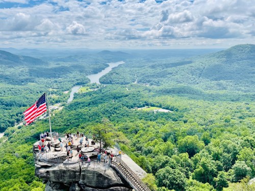 A panoramic view from a rocky overlook with a large American flag, surrounded by lush green mountains and a winding river.