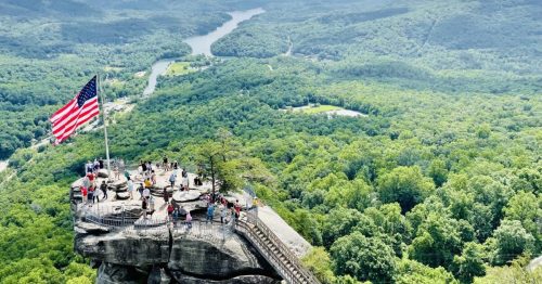 A panoramic view from a rocky overlook with an American flag, surrounded by lush green mountains and a winding river below.