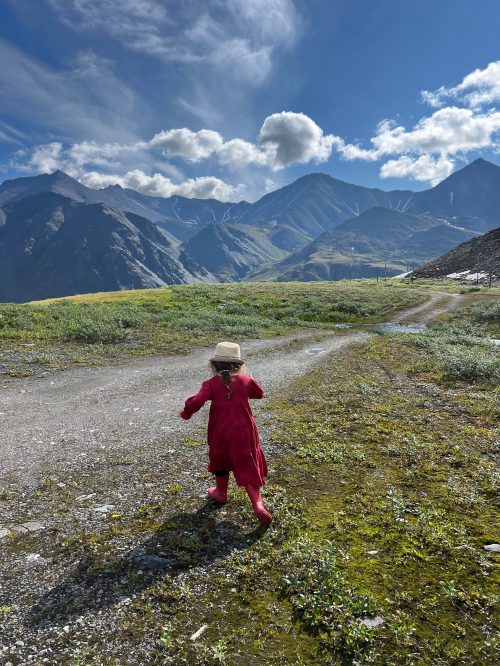 A child in a red dress and hat runs along a gravel path, surrounded by mountains and a blue sky with clouds.