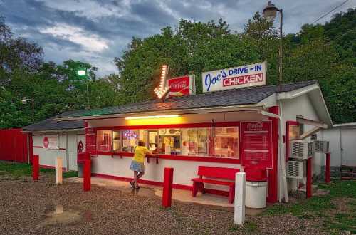 A retro drive-in restaurant with a bright sign, featuring a customer ordering at the window under a cloudy sky.