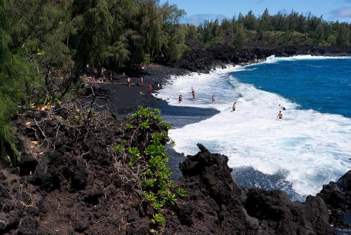 A scenic view of a black sand beach with people swimming, surrounded by lush greenery and rocky shoreline.