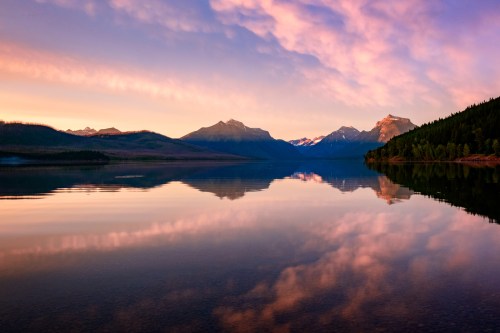 Serene lake at sunrise, reflecting mountains and colorful clouds in calm waters.