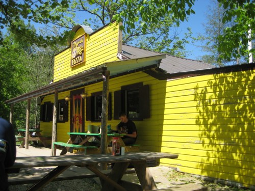 A bright yellow pizza shop surrounded by trees, featuring outdoor seating and a sunny blue sky.