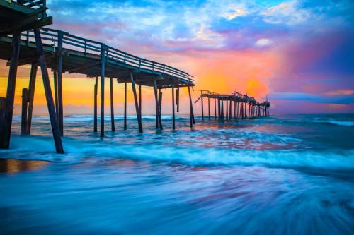 A colorful sunset over a wooden pier extending into the ocean, with waves gently lapping at the shore.