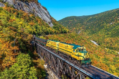 A yellow and green train travels along a trestle bridge surrounded by vibrant autumn foliage and mountains.