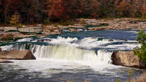 A scenic waterfall cascades over rocks, surrounded by autumn foliage and a tranquil river.